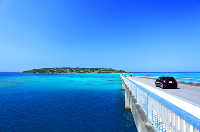 澄み切った青い空の下、エメラルドグリーンの美しい海に架かる長い橋を黒い車が走っています。遠くには緑豊かな島が見え、爽快なドライブを楽しむ風景です。