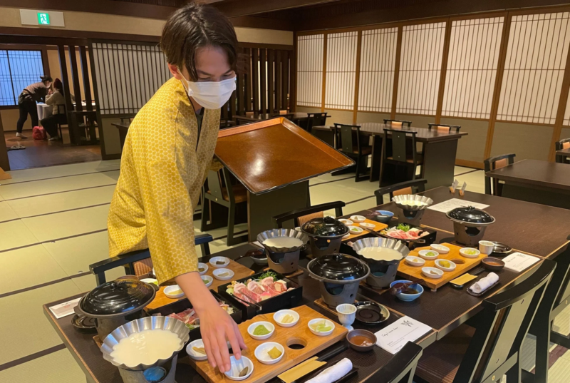 和風の個室で、マスクを着用した従業員が会席料理の準備をしている様子です。