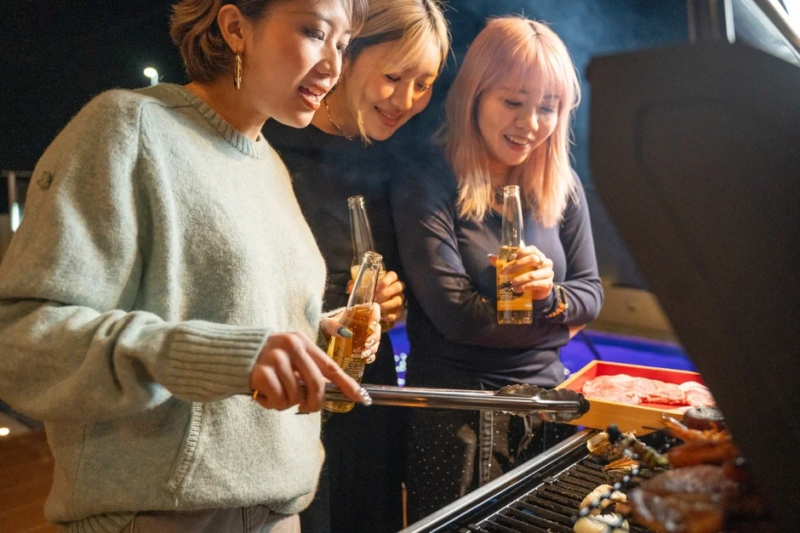 夜、3人の女性が屋外でバーベキューを楽しんでいる様子です。皆ビール瓶を片手に笑顔で、グリルには肉やシーフードが焼かれています。和やかな雰囲気の集まりです。