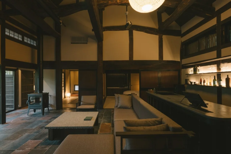 伝統的な梁が見える日本家屋の広い空間に、モダンなソファやバーカウンター、薪ストーブが配されたラウンジ