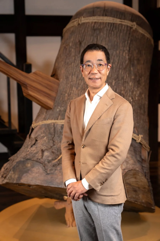 笑顔の男性が、大きな木製の伝統的なオブジェを背景に立っている