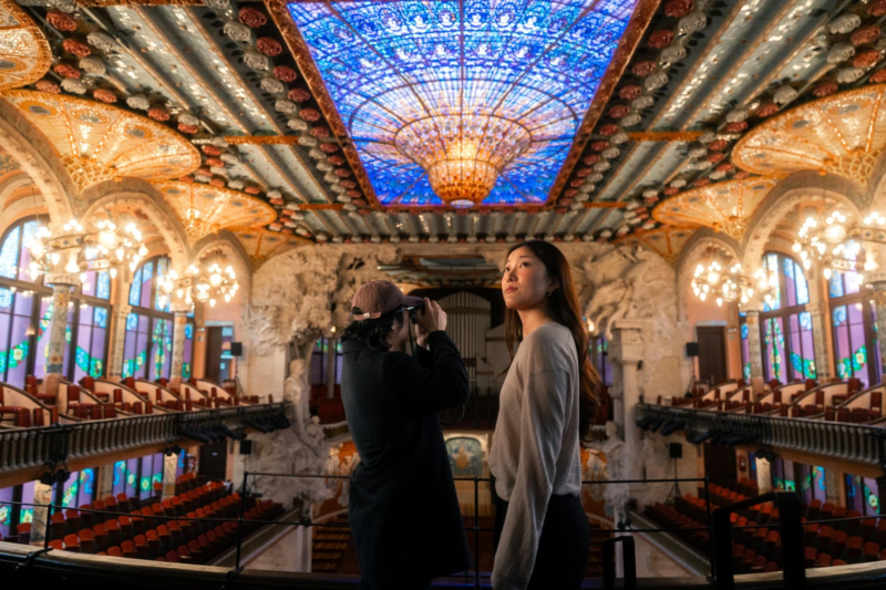 豪華絢爛なコンサートホールの内部で、一人の人物がカメラで撮影し、もう一人が壮麗なステンドグラスの天井を見上げています