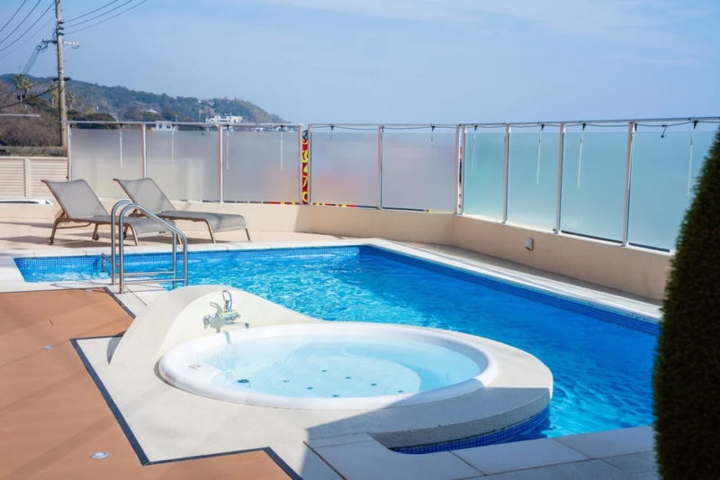 晴れた日の屋外に広がる青いプールとジャグジーの光景。プールサイドにはデッキチェアが2つ並び、リラックスできる空間が広がっています。遠くには山々が見え、穏やかなリゾートの雰囲気が感じられます。