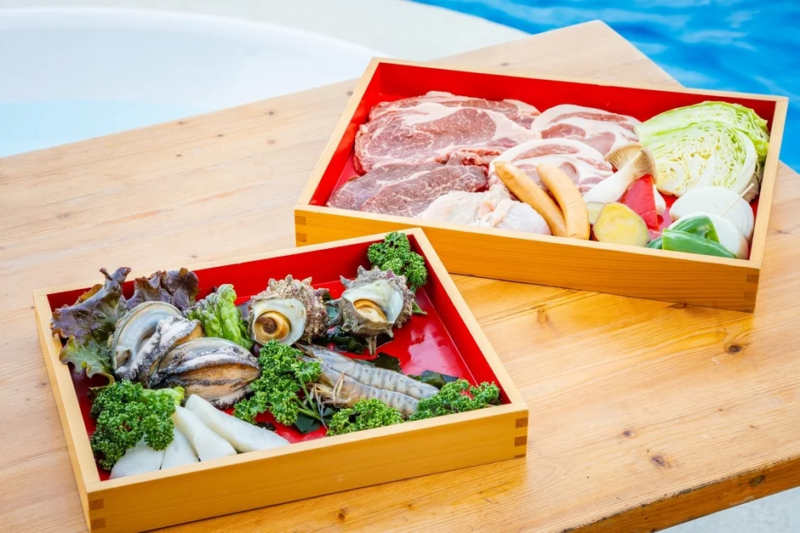 木製トレーに盛り付けられた豪華なバーベキュー食材のセット。牛肉、豚肉、鶏肉、ソーセージ、野菜に加え、アワビ、サザエ、エビ、イカなどの新鮮な海鮮が豊富に用意されており、屋外での贅沢な食卓を演出します。
