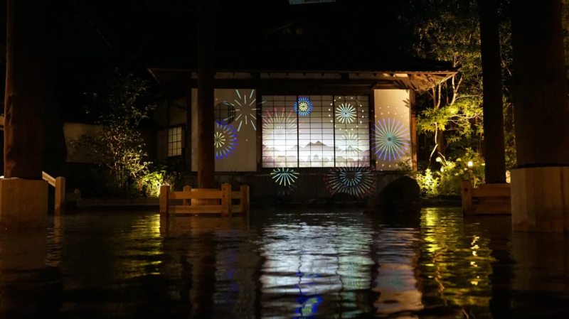 夜の日本家屋にプロジェクションマッピングで花火が投影され、水面にその光が幻想的に反射している。和の雰囲気。