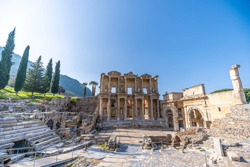 トルコのエフェソスにあるケルスス図書館の壮大な古代遺跡の全景です。青い空の下、石造りの図書館のファサードが堂々とそびえ、周囲には他の遺跡や緑豊かな木々、山々が広がっています。