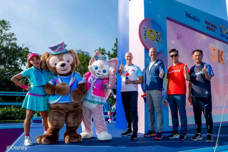ディズニーキャラクターのダッフィーとリーナベル、キャスト、そして男性たちがAIA主催のランニングイベントで笑顔でポーズをとっている様子。10Kや3Kの表示があり、イベントの活気ある雰囲気が伝わる一枚。