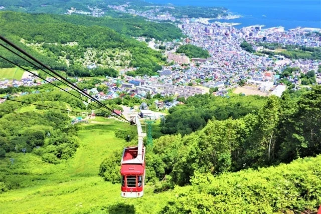 山の斜面を走る赤いゴンドラと、その先に広がる美しい街並み、そして青い海を一望できる、緑豊かな夏の絶景
