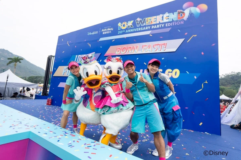 香港ディズニーランドで開催された「10K WEEKEND 20TH ANNIVERSARY PARTY EDITION」イベントで、ドナルドダックとデイジーダックが参加者たちと楽しそうにポーズをとっている。カラフルな衣装と紙吹雪が祝祭的な雰囲気を演出している。