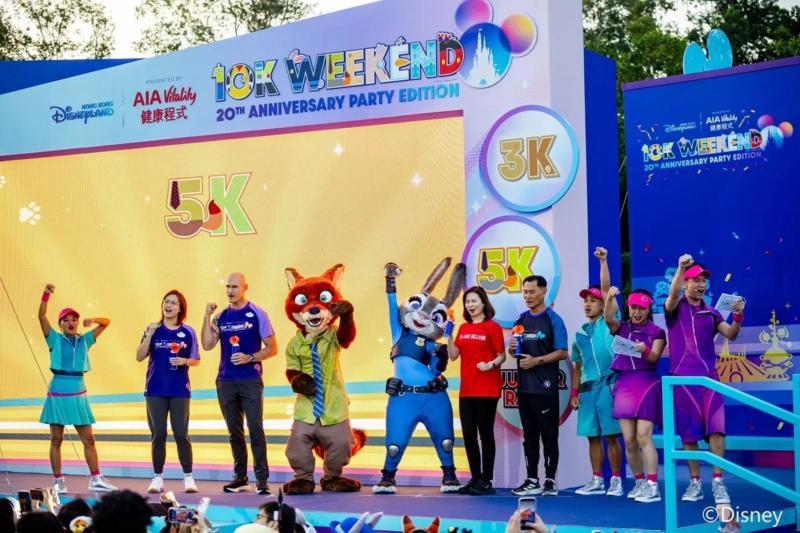 香港ディズニーランドで開催された「10K WEEKEND 20TH ANNIVERSARY PARTY EDITION」のステージイベントの様子。ズートピアのニックとジュディのキャラクターが、MCや参加者らしき人々とともにポーズをとっており、背景には「5K」や「3K」の表示が見える。祝祭的な雰囲気で、イベントの盛り上がりが伝わります。