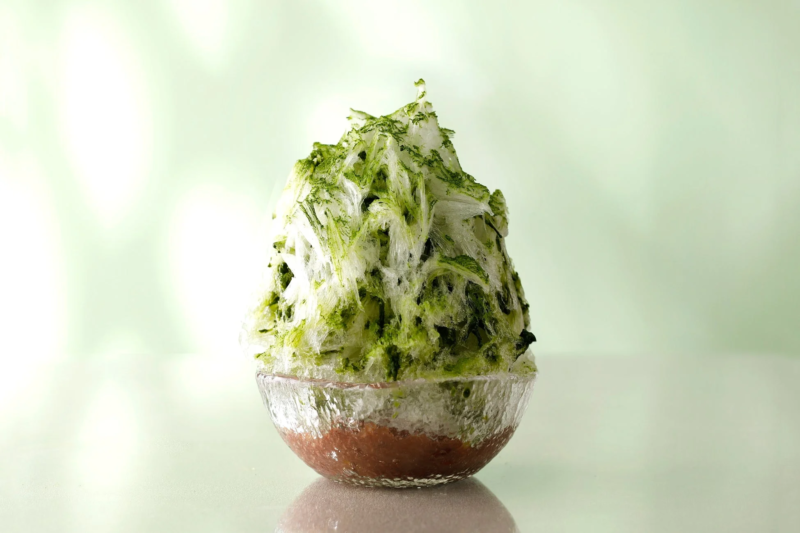抹茶の緑と小豆の茶色が鮮やかな、透明な器に入った宇治金時のかき氷です。ふんわりと盛られた氷が清涼感を誘い、夏の暑い日にぴったりの和風デザートを表しています。