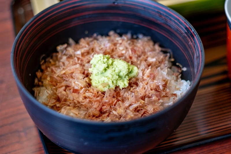 鰹節がたっぷり乗ったご飯にワサビが添えられた和食のどんぶりのクローズアップ。木製のトレイに置かれ、温かみのある食卓の風景を写しています。