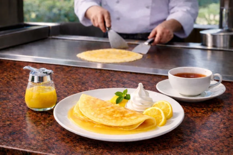 シェフが鉄板でクレープを調理しており、手前にはレモンとクリームが添えられた美味しそうなクレープと紅茶が並べられています。