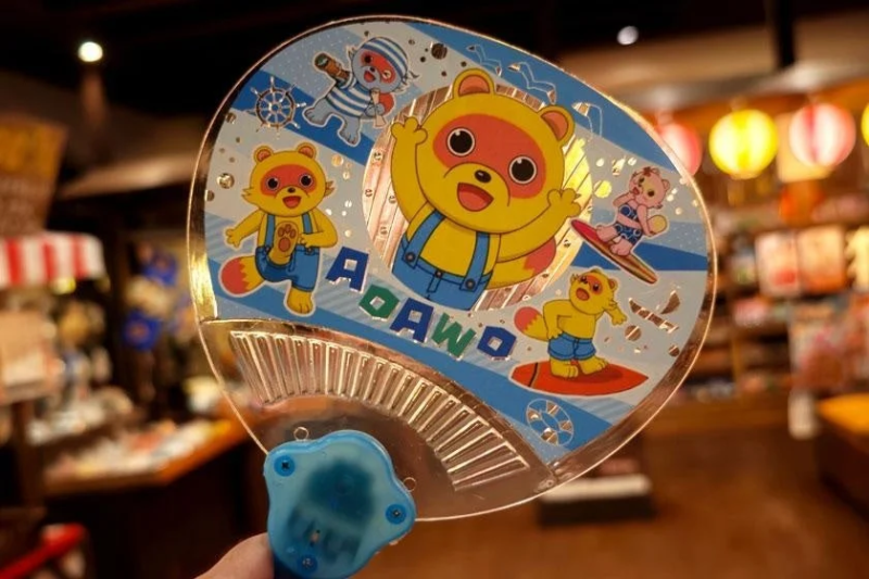 透明なうちわに、アライグマ風の可愛いキャラクターたちがマリンスポーツを楽しむ様子が描かれています
