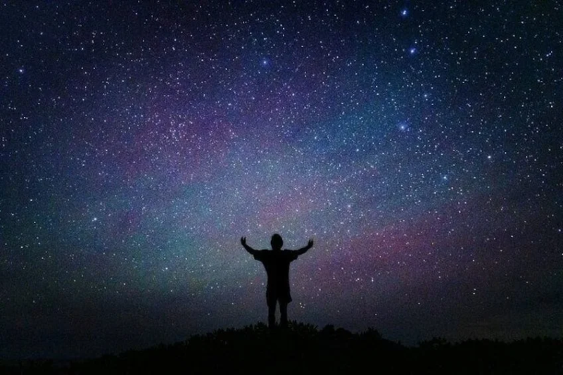 無数の星が輝く夜空の下、丘の上に立つ人物のシルエットが印象的な画像です。両腕を広げ、満天の星を見上げている姿は、宇宙の広がりと神秘を感じさせます。