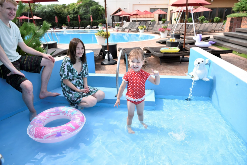 リゾートホテルのプールサイドで、幼い女の子が水遊びを楽しんでおり、そのそばで両親らしき男女が笑顔で見守っています