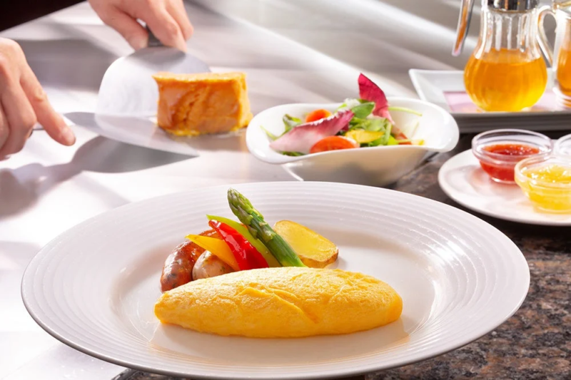 ホテルビュッフェのような場所で提供される豪華な朝食の様子です。手前にはふわふわのオムレツ、ソーセージ、野菜が盛り付けられたプレートがあり、奥では別の料理が取り分けられています。