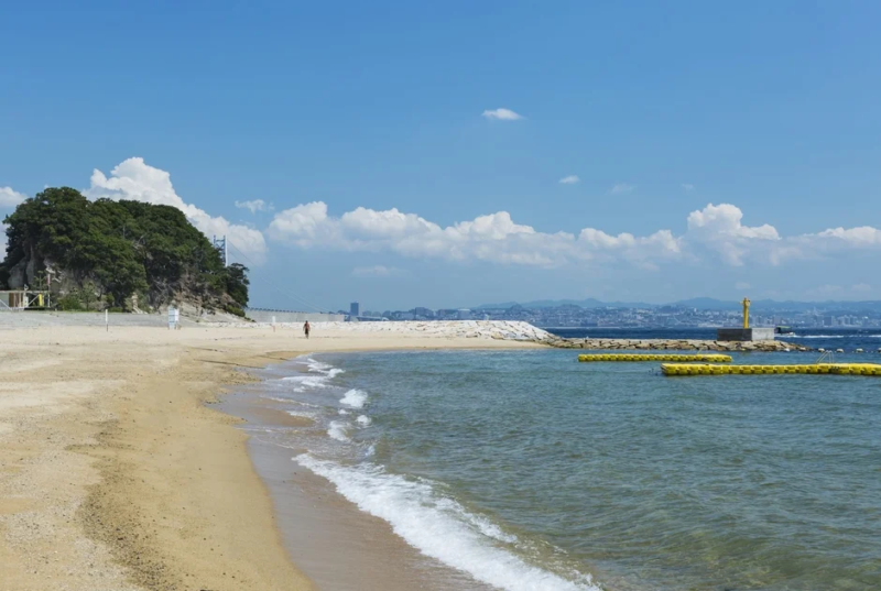 晴れた夏の日に広がる砂浜と青い海、遠くには吊り橋が見える