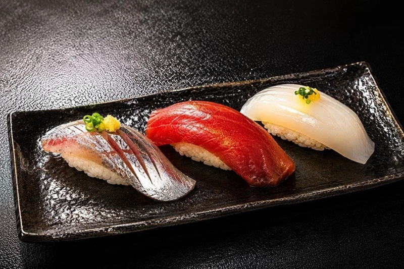 黒い長方形の皿に、赤身のマグロ、光り物の魚、白いイカの3種類の握り寿司が美しく盛り付けられています。