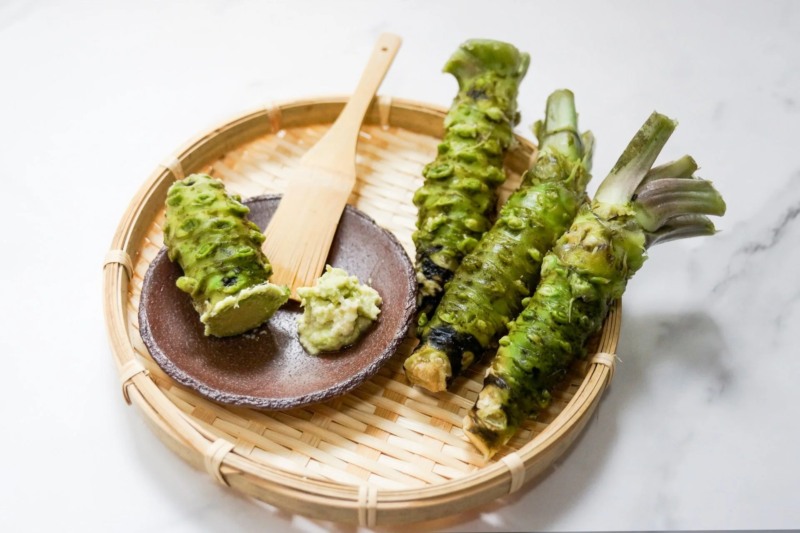 新鮮な本わさびの根茎が数本、竹ざるに盛られています。一本はすりおろされており、小皿に盛られたわさびと木製のヘラが添えられています。和食に欠かせない薬味の準備風景です。