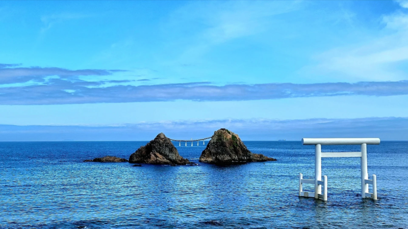 青い海と空が広がる穏やかな風景で、海中に立つ白い鳥居と、注連縄で結ばれた二つの岩（夫婦岩）が特徴的です