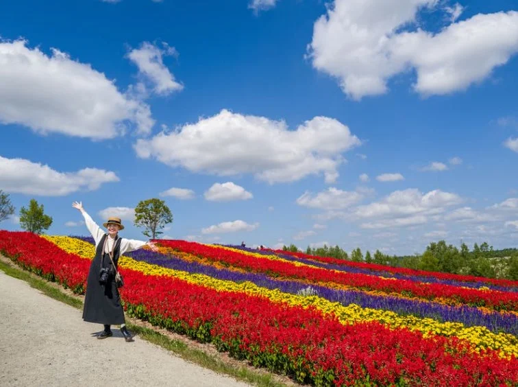 麦わら帽子をかぶった女性が、赤、黄、紫のカラフルなストライプ状の花畑で両腕を広げて笑顔を見せている。青空と白い雲が広がる丘陵地帯の美しい風景。
