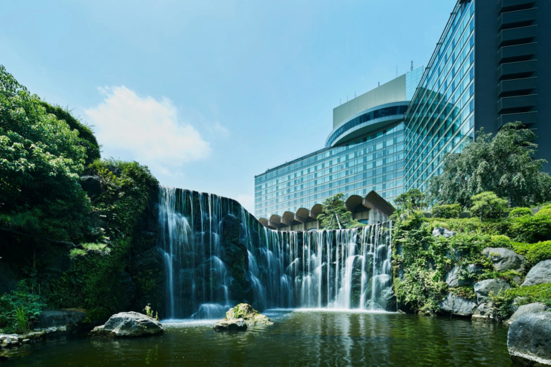 近代的な建物と豊かな緑に囲まれた美しい滝と池のある庭園の風景です。青空の下、勢いよく水が流れ落ちる様子が清々しい印象を与えています。