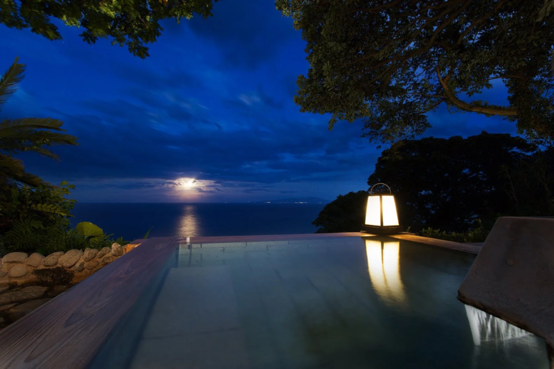 月明かりが海面に反射する夜、広大な海を望む露天風呂の情景。湯船の傍らには灯籠が優しく光り、幻想的で心安らぐ空間が広がっています。