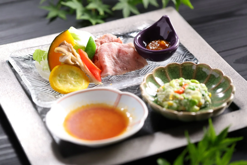 透明なガラスの皿に盛り付けられた薄切りの肉と色鮮やかな野菜、そして二つの小鉢に入ったソースと和え物が並ぶ、上品な和食の膳です。
